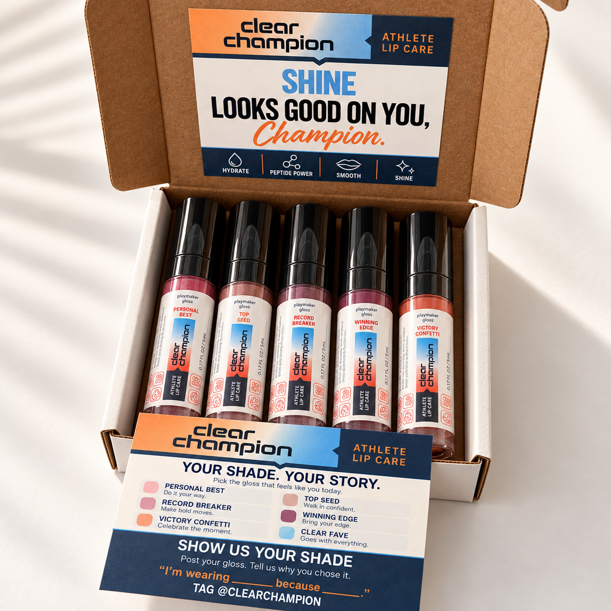 An open cardboard box of Clear Champion Athlete Lip Care lip tint tubes, with a branded insert card placed in front. The box is photographed on a white surface with soft diagonal shadows.