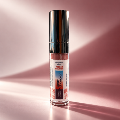 A clear cylindrical gloss bottle with a reflective silver cap, standing upright on a pink studio surface. The front label reads 'shaymaker gloss' and 'Record Breaker Clear Champion'.
