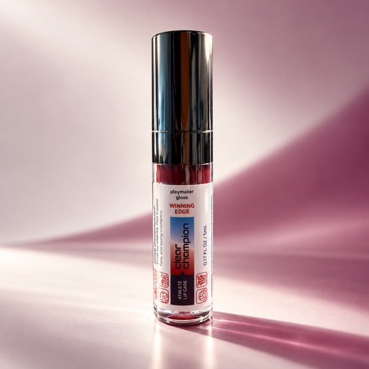 A single bottle of Playmaker Gloss Winning Edge lip color in a clear cylindrical glass bottle with a reflective silver cap, standing on a pink surface with soft pink shadows.